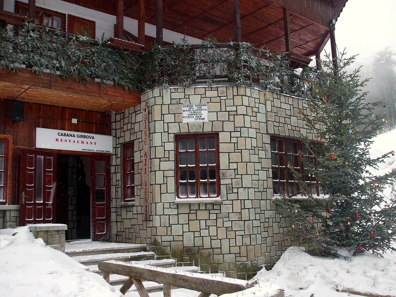 Colțul de piatră al cabanei iarna, cu firma restaurantului, panoul vertical de întâmpinare și plăcuța de la intrare.