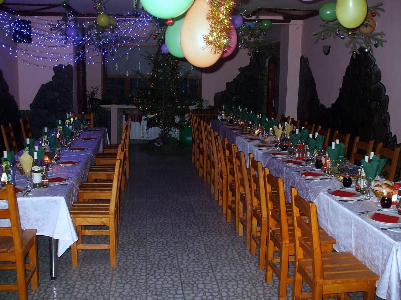 Mese lungi puse pentru o petrecere, cu fețe de masă albe, șervete verzi împăturite, lumânări și carafe cu vin.